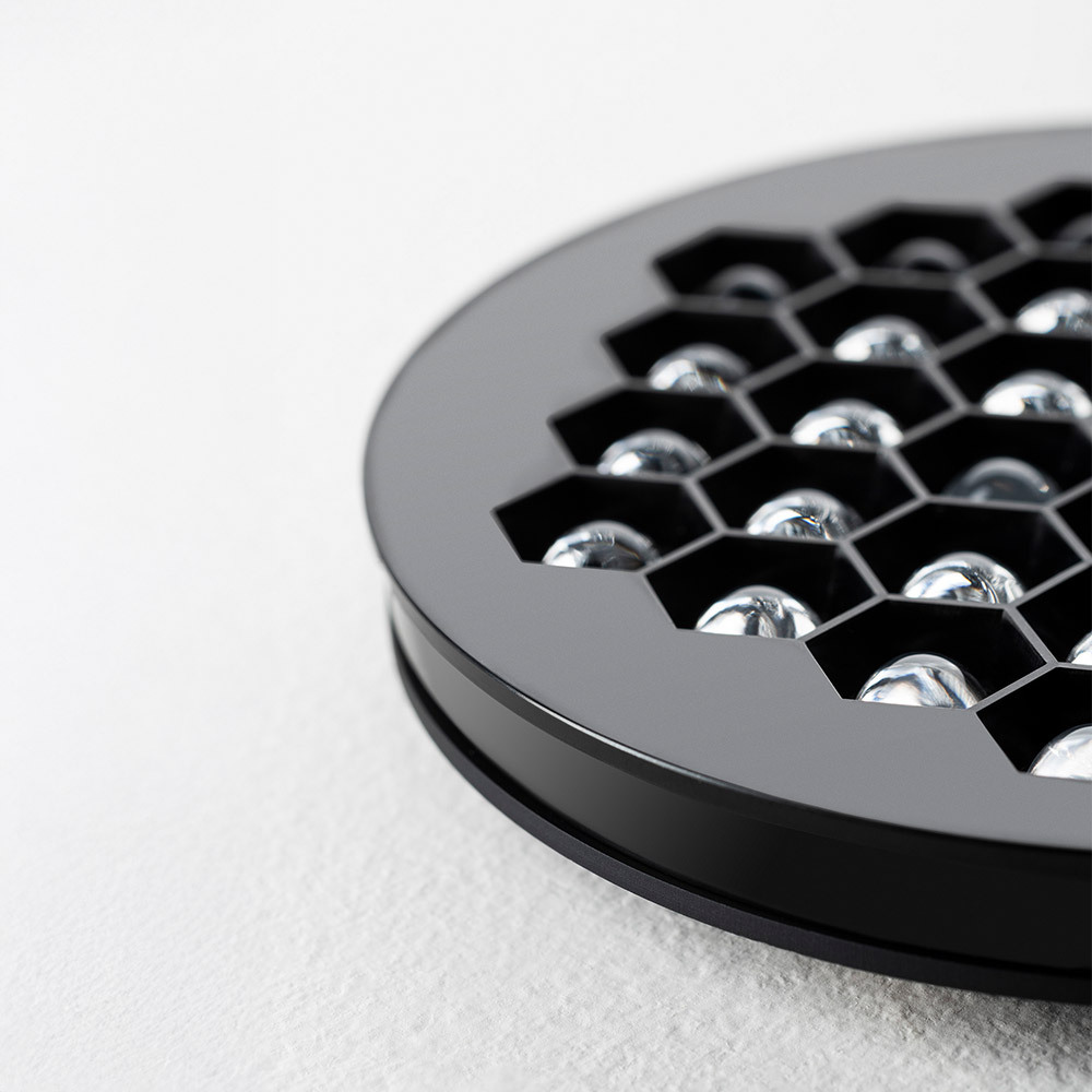 Close-up of an Artemide lighting module with a circular design, featuring a black structure and a top surface characterized by a hexagonal grid housing transparent optical lenses. The image highlights the precision craftsmanship and high-quality materials used.