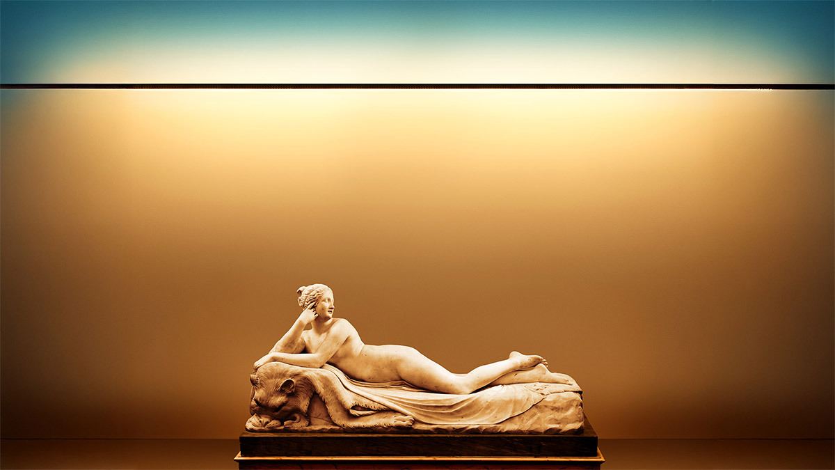 Classical statue of a reclining woman with a lion at her feet, lit by an Artemide light gradient fading from warm tones to blue on a neutral wall.