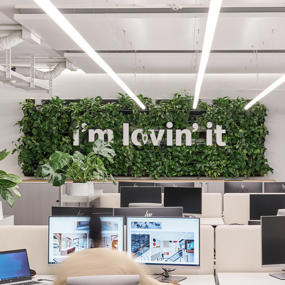 Green wall with “i’m lovin’ it” sign in a modern office with plants and workstations.