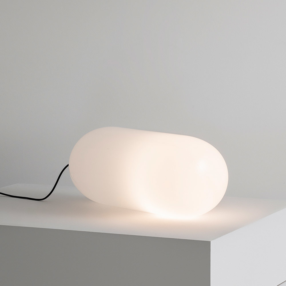 Lampe de table Artemide en verre blanc dépoli en forme de pilule diffusant une lumière douce et chaude, posée sur surface blanche avec câble noir