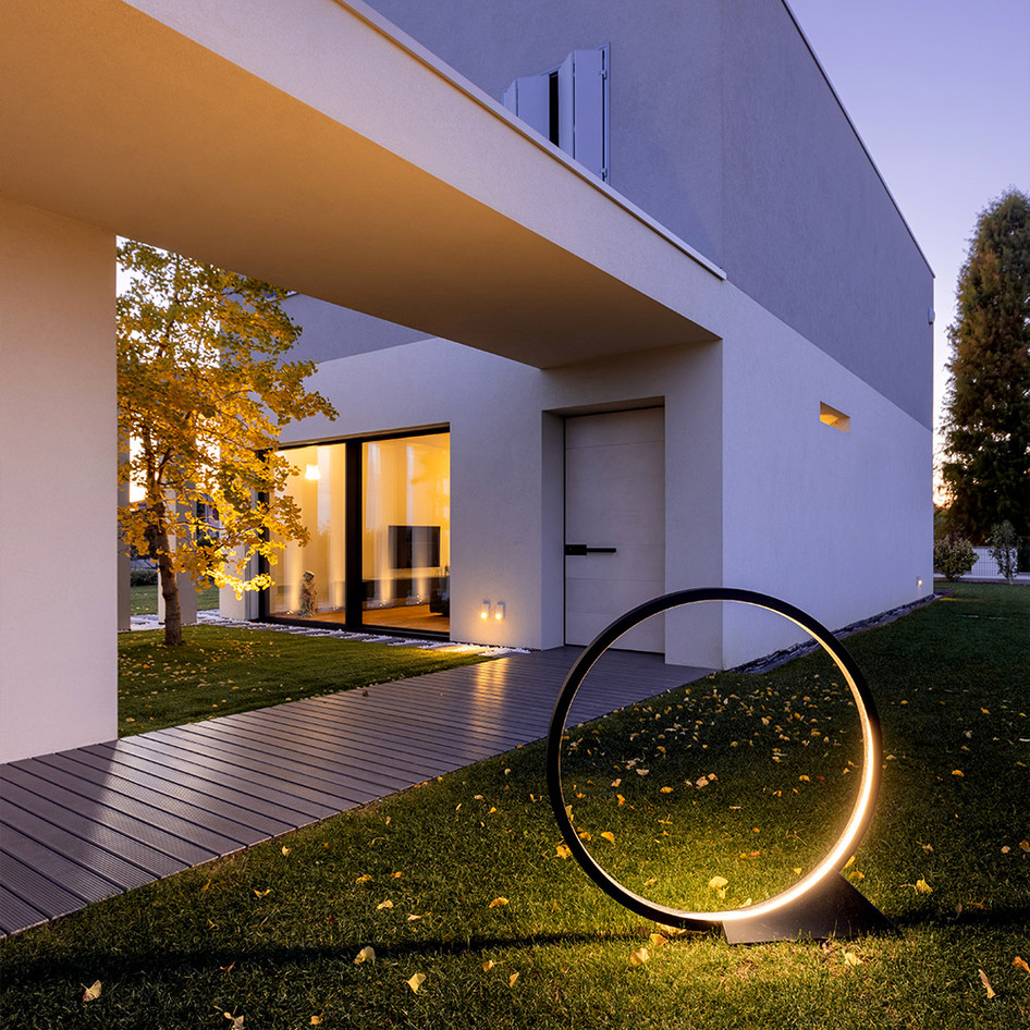 Lampadaire LED circulaire Artemide sur pelouse au crépuscule, diffusant une lumière chaude près de l'entrée d'une maison blanche minimaliste