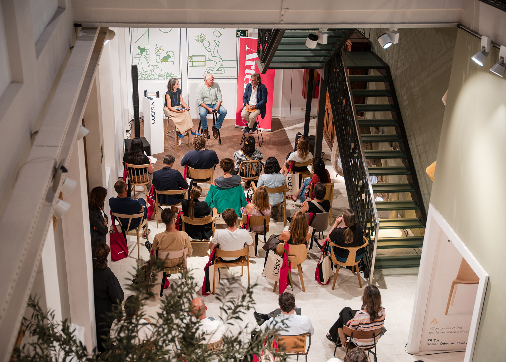 Aerial view of a panel discussion with three speakers facing an audience seated on wooden chairs in a bright interior space.