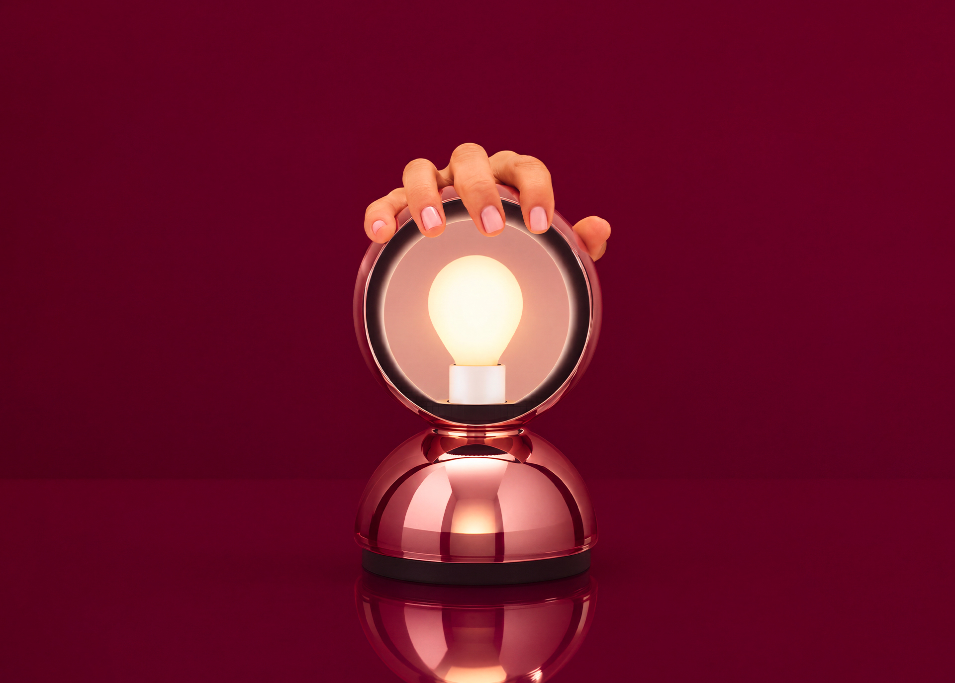 Mano tocando una lámpara de mesa oro rosa con marco circular espejado y bombilla Edison encendida sobre superficie reflectante, fondo rojo