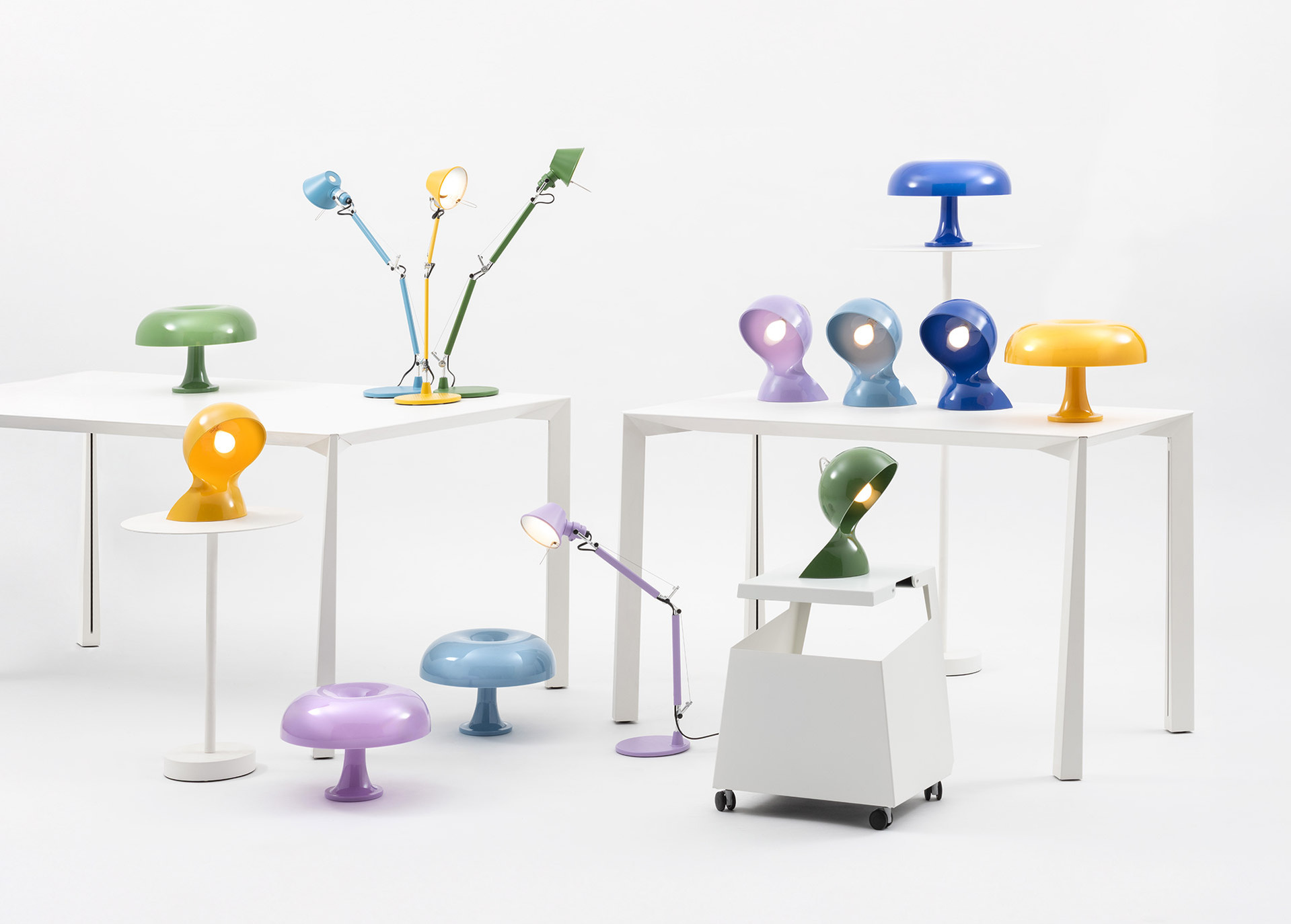 Composition of Dalù, Nessino and Tolomeo lamps in various colors, arranged on white tables against a neutral background.
