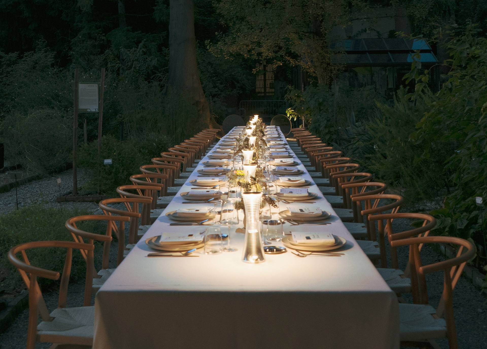 Long outdoor table in an evening garden, with wooden chairs, elegant plates, and 'Come Together' lamps placed on the table.