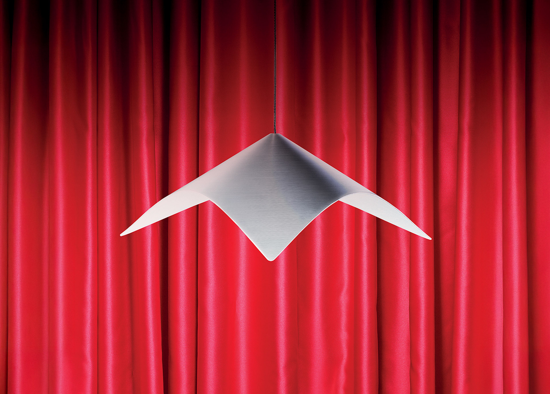 Iperbole pendant lamp in satin-finished metal, with a geometric form, suspended in front of a red curtain. Photo by Pierpaolo Ferrari.