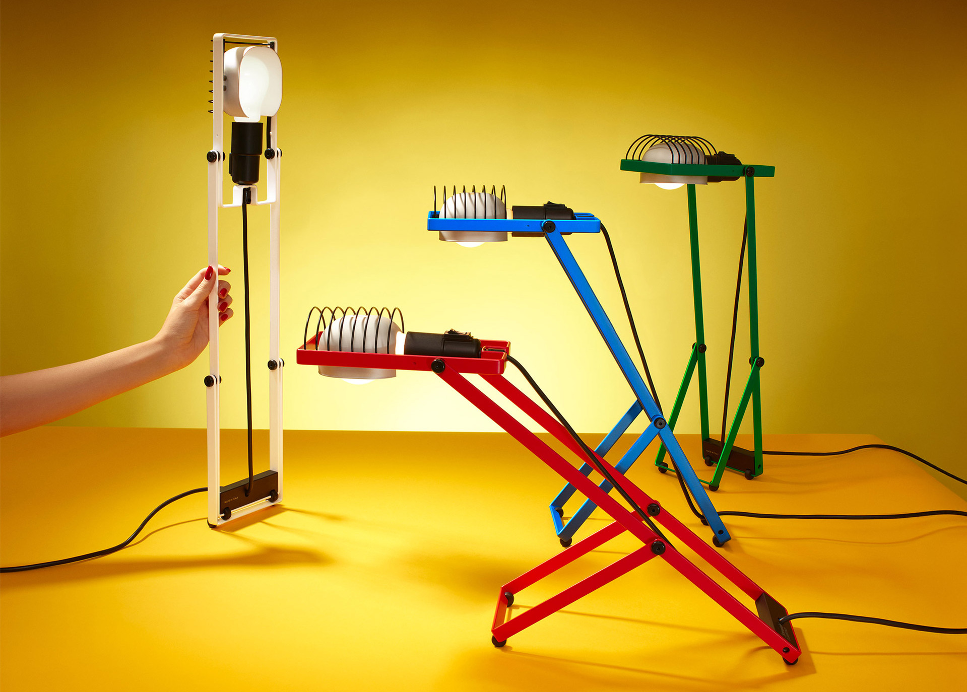 Four Sintesi lamps in white, red, blue, and green arranged in different configurations against a yellow background, photographed by Pierpaolo Ferrari.