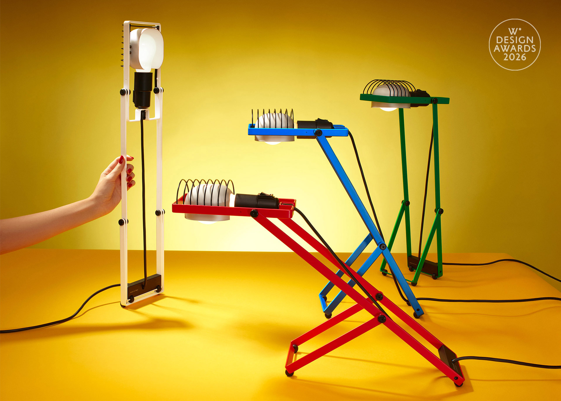 Four Sintesi lamps in white, red, blue, and green arranged in different configurations against a yellow background, photographed by Pierpaolo Ferrari, featuring the Wallpaper* Design Awards logo.