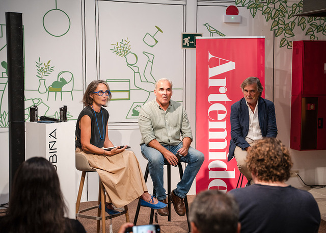 Three speakers at an Artemide panel discussion, seated before a red Artemide banner and illustrated wall, addressing an audience.