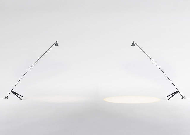Dos lámparas de pie minimalistas oscuras con largos brazos inclinados y pantallas cónicas, proyectando haces de luz concentrados sobre una superficie blanca.
