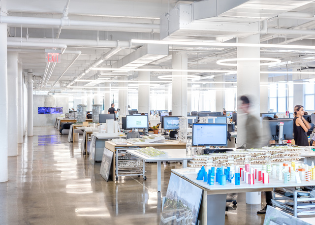 BIG’s New York offices in a large open-plan workspace with desks, computers, architectural models, and linear ceiling lighting.