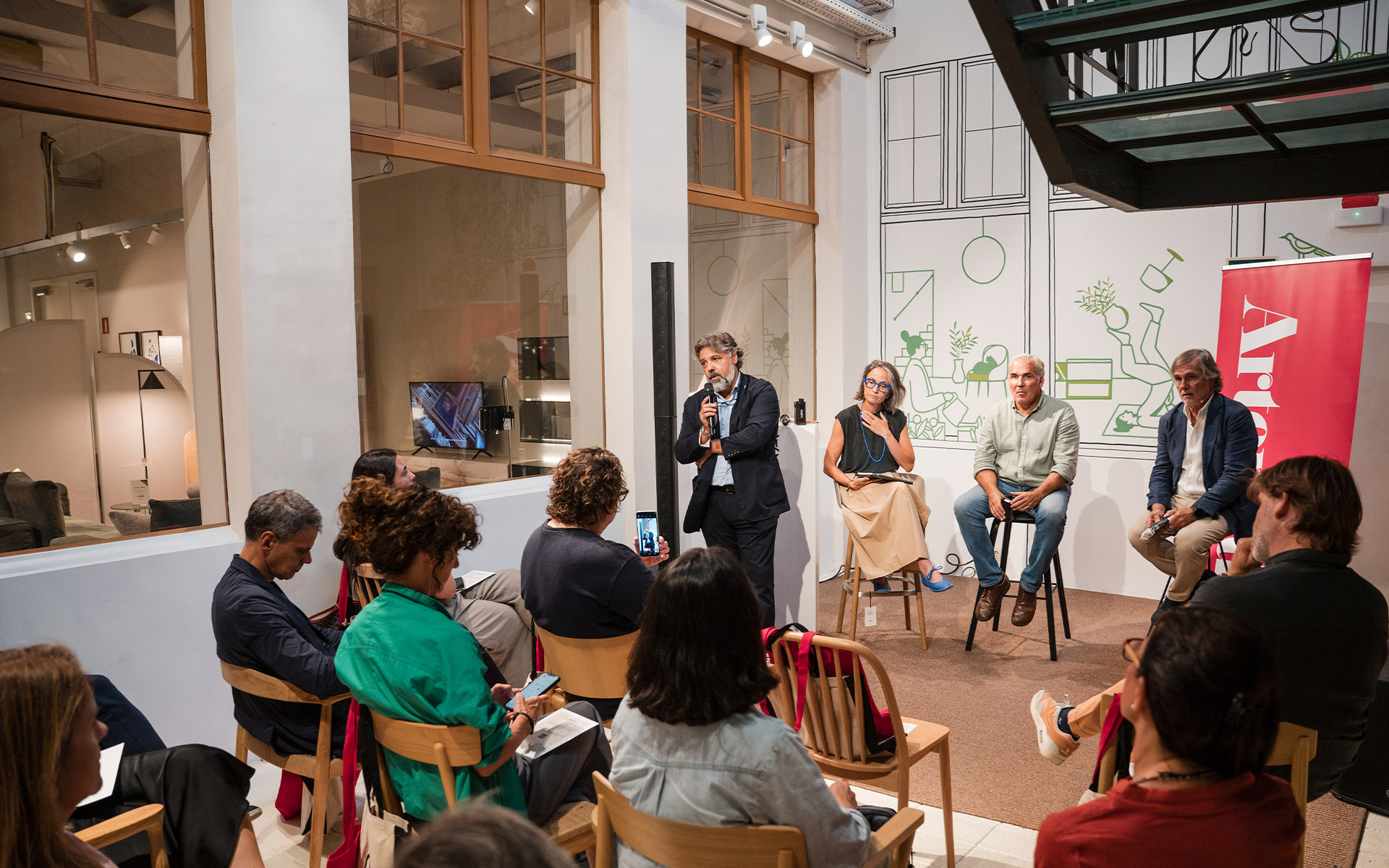 Panel discussion with four speakers on stools facing an audience in a bright gallery space with illustrated wall murals.