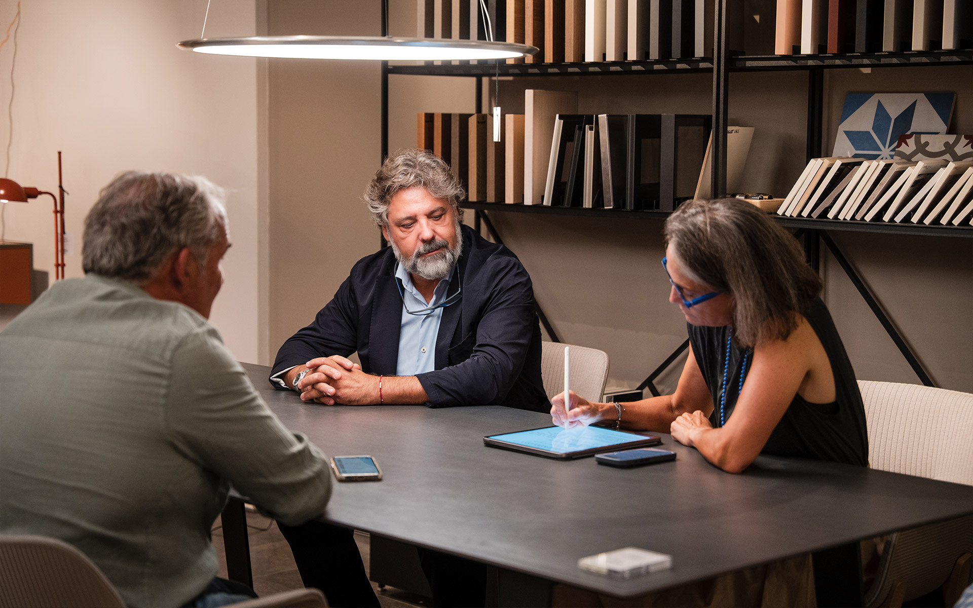 Three professionals in a meeting around a dark table, illuminated by an Artemide pendant lamp, reviewing project on a tablet.
