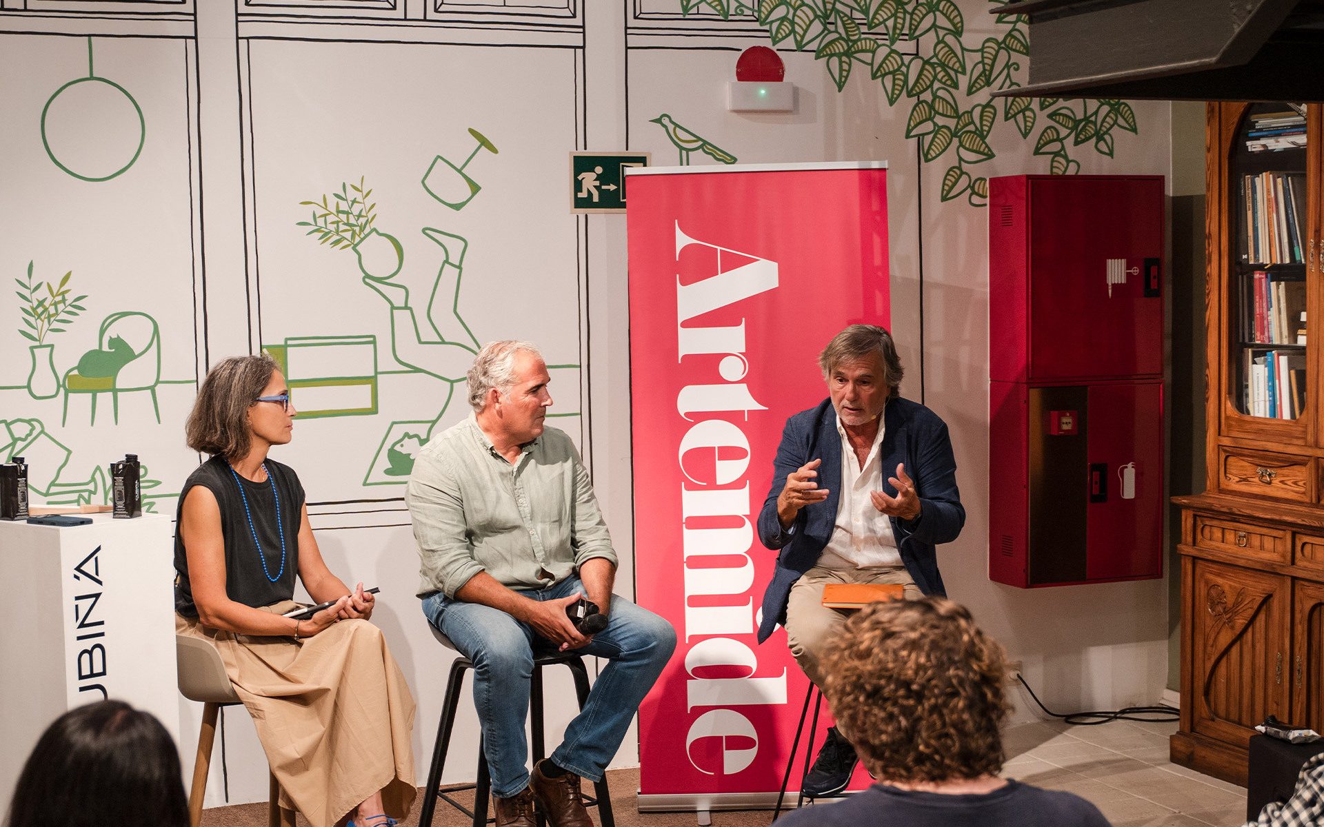 Three people seated in conversation at an Artemide brand event, with a red Artemide banner visible in the background.