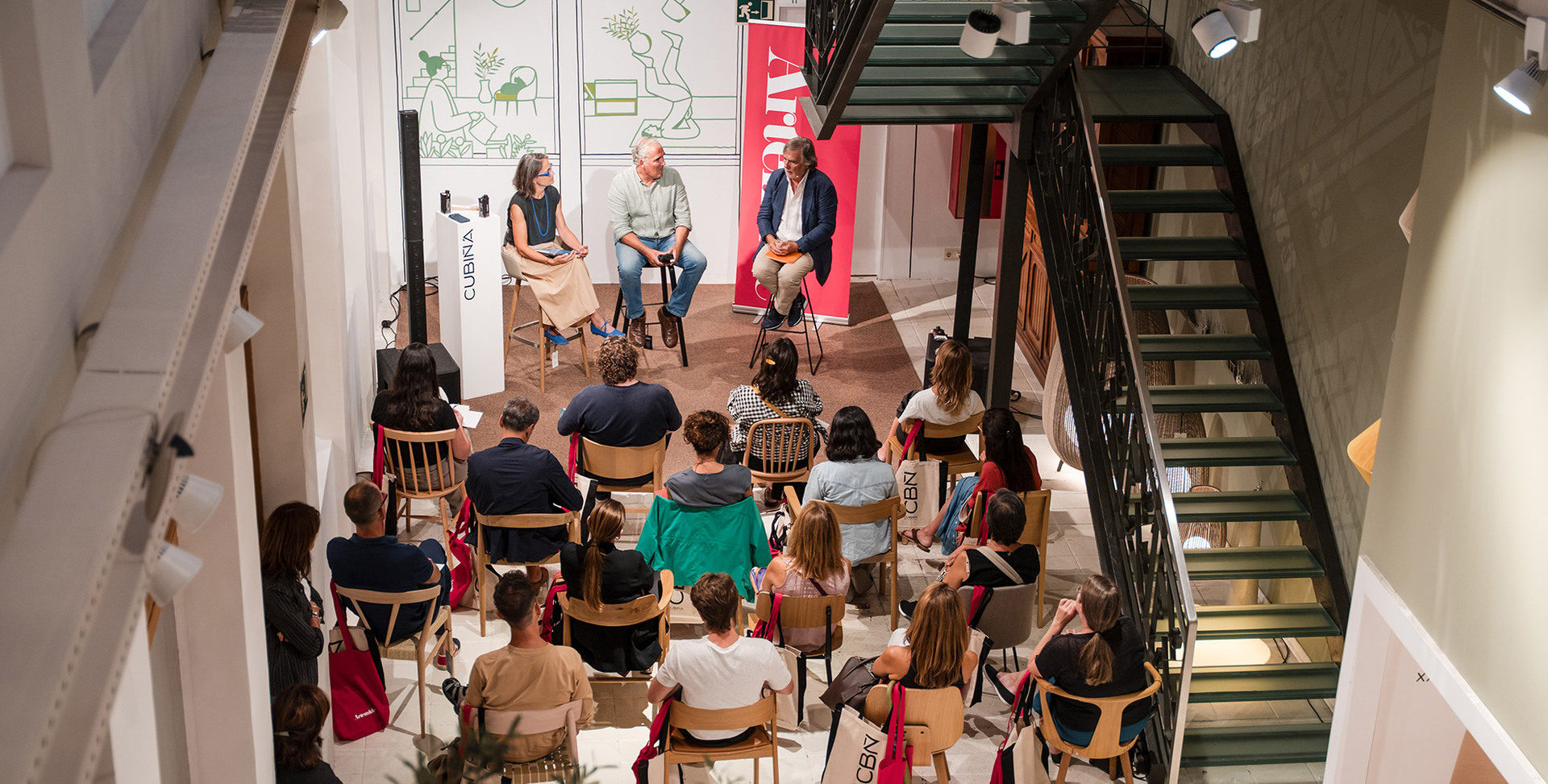 Aerial view of a panel discussion with three speakers facing an audience of about 20 people seated on wooden chairs in a modern venue.