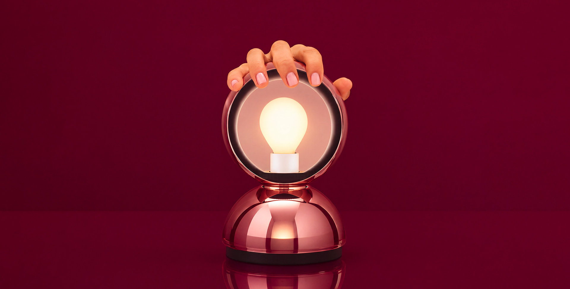 Mano tocando una bombilla encendida dentro de una lámpara de mesa esférica de metal oro rosa con base cromada redondeada.