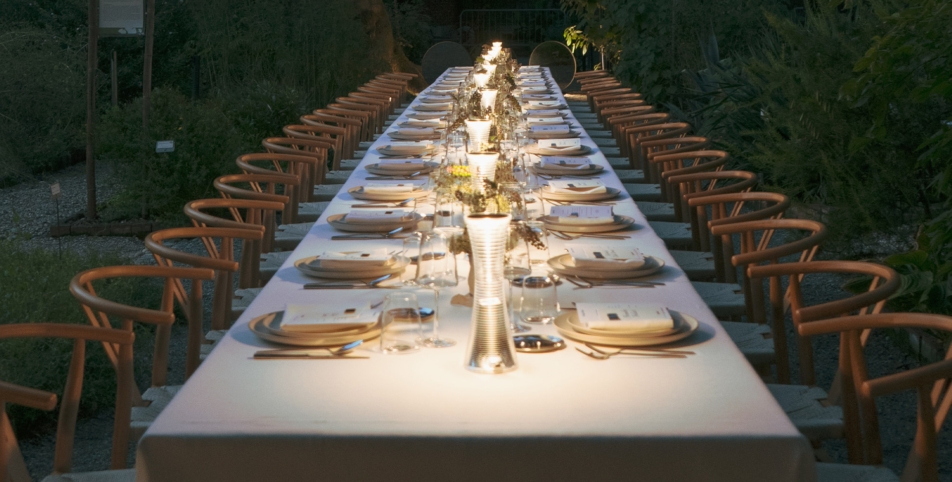 Long outdoor table in an evening garden, with wooden chairs, elegant plates, and 'Come Together' lamps placed on the table