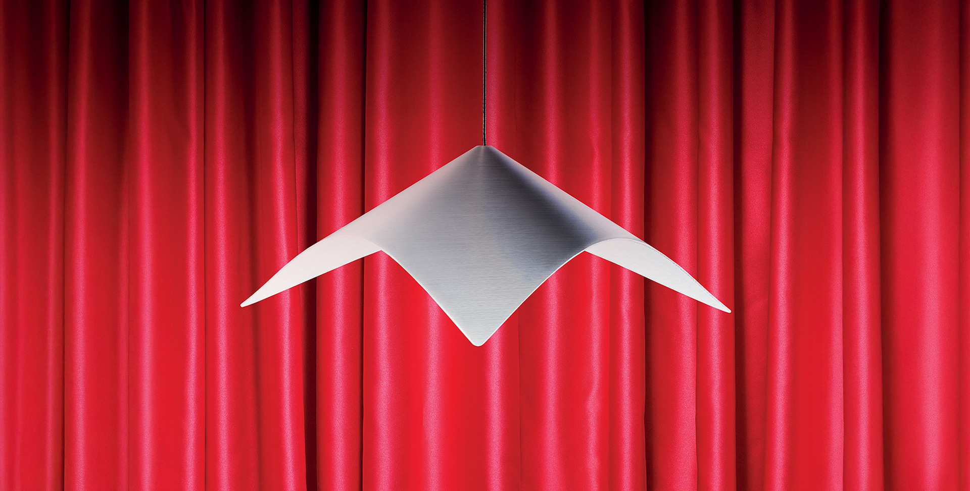 Iperbole pendant lamp in satin-finished metal, with a geometric form, suspended in front of a red curtain. Photo by Pierpaolo Ferrari.