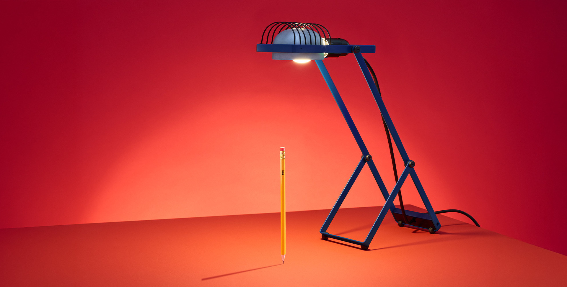 Blue Sintesi lamp illuminating a vertical pencil against a red background, photographed by Pierpaolo Ferrari.