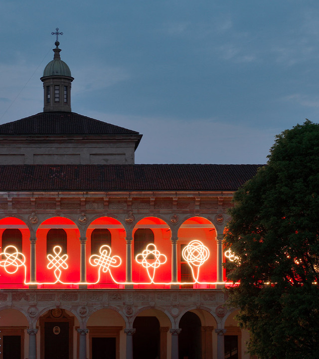 Facade of the courtyard of the University of Milan at dusk, with a linear light installation drawing interwoven motifs across the arches.