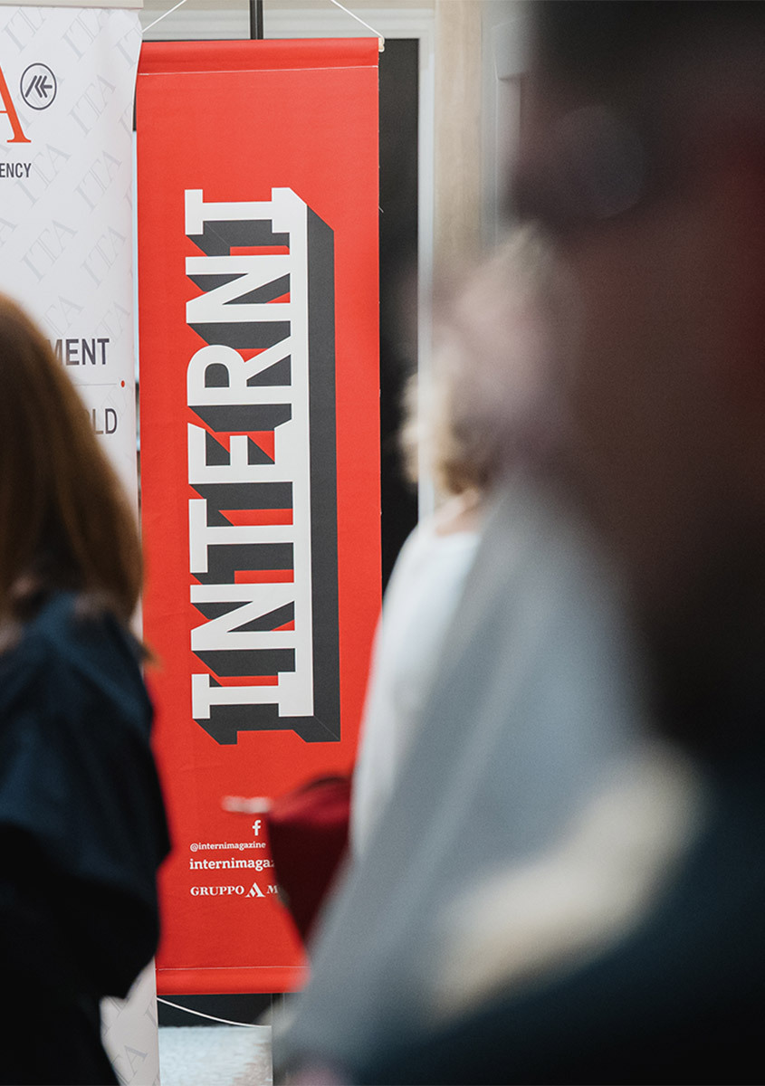 Striscione verticale rosso con il testo in grassetto 'INTERNI' esposto a un evento di design, con visitatori sfocati in primo piano.