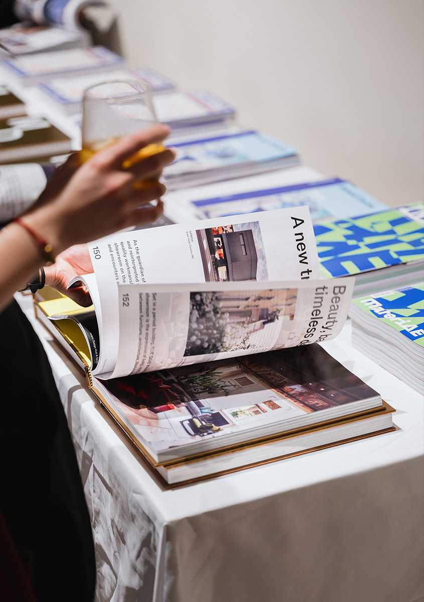 Persona che sfoglia un catalogo a un evento, con un bicchiere di vino in mano mentre scorre le pagine con prodotti di illuminazione.