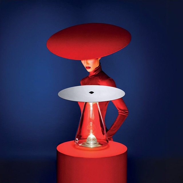 Lámpara de mesa Artemide con base cónica transparente y pantalla de disco plano sobre pedestal rojo, estilizada con modelo en rojo con sombrero de ala ancha
