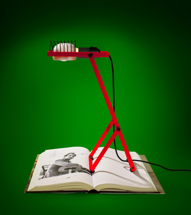 Red Sintesi lamp placed over an open catalog showing a black-and-white photo of Ernesto Gismondi, against a green background, photographed by Pierpaolo Ferrari.