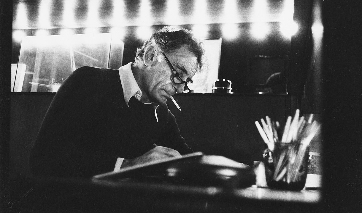 Historic photo of Gianfranco Frattini working at his desk, in a black-and-white portrait lit by a row of lights in the background.