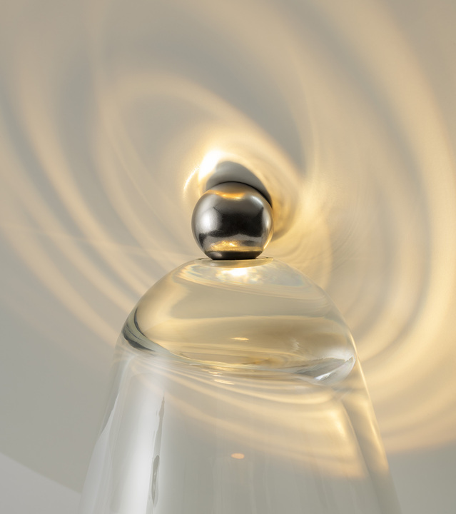 Primer plano de la parte superior de una lámpara de vidrio transparente con remate esférico cromado, proyectando ondas de luz concéntricas en el techo