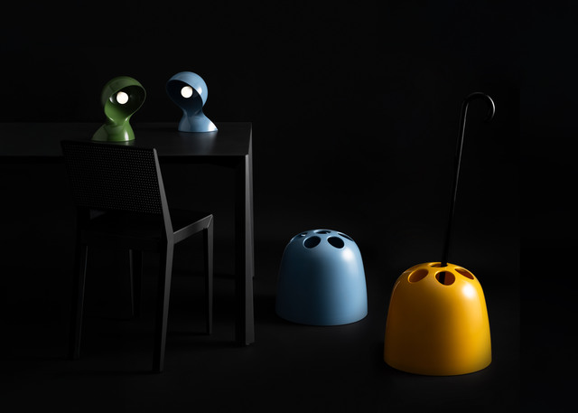 Composition with two Dalù lamps, one green and one light blue, placed on a table and accompanied on the floor by two Dedalo umbrella stands against a black background.