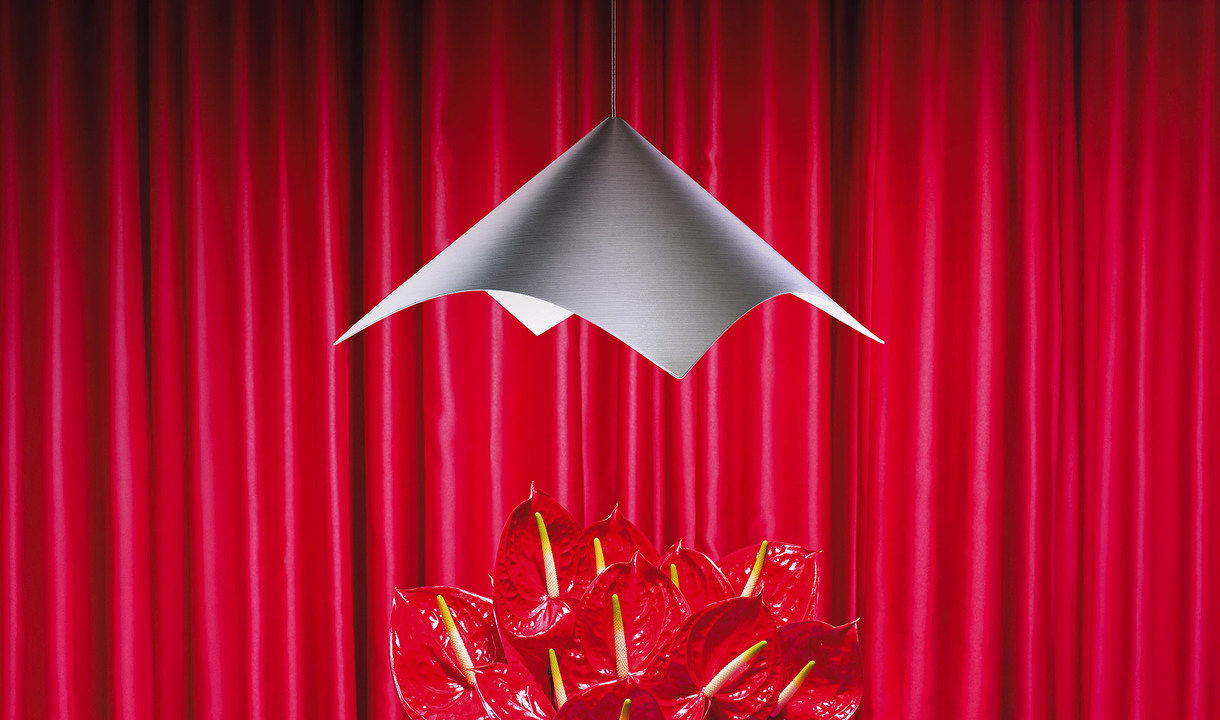 Iperbole metal pendant lamp with a sculptural shape, suspended above a group of red anthuriums in front of a backdrop of red curtains. Photo by Pierpaolo Ferrari.