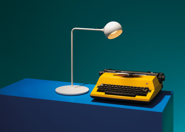 Immagine della lampada Ixa bianca su un tavolo blu e con affianco una macchina da scrivere gialla.