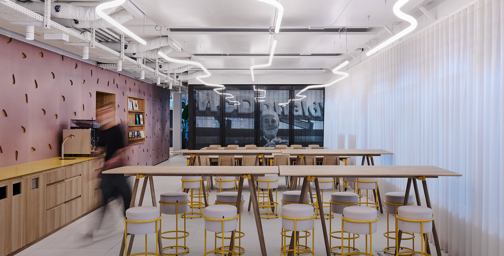 Corporate kitchen area with high tables, yellow stools and curved LED lights on the ceiling.