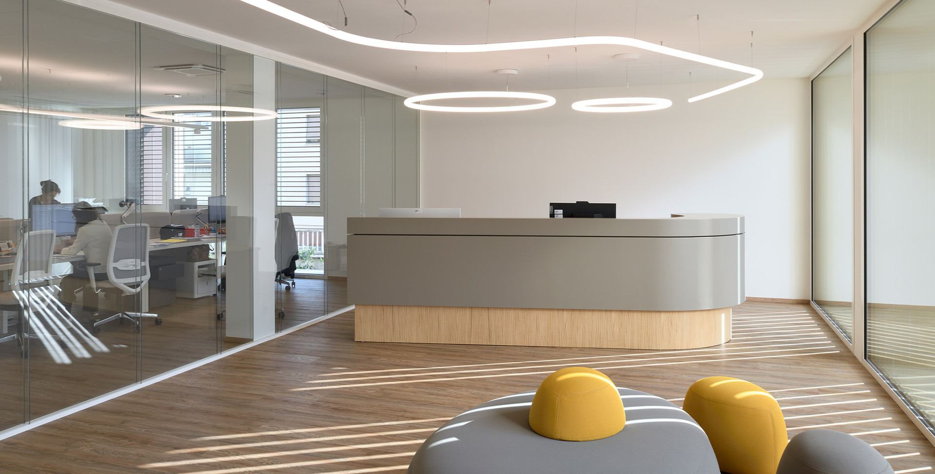 Reception area of the new Fornaroli Polymers headquarters with a curved desk, wooden flooring and glass walls, illuminated by the Alphabet of Light suspension system.