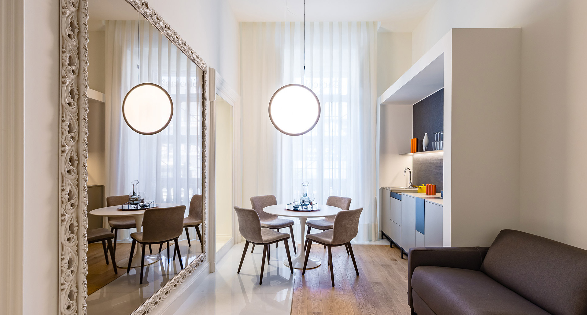 Elegant dining area with two Artemide circular pendant lights above a round white table with taupe chairs, sheer curtains, and a large ornate mirror.