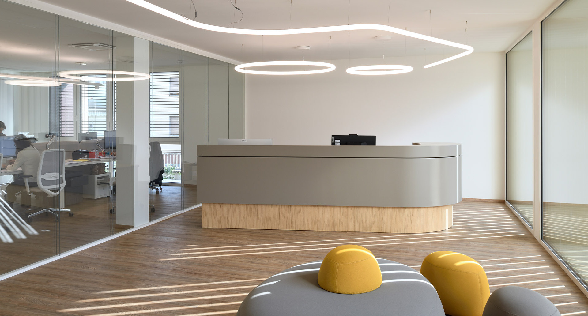 Reception area of the new Fornaroli Polymers headquarters with a curved desk, wooden flooring and glass walls, illuminated by the Alphabet of Light suspension system.