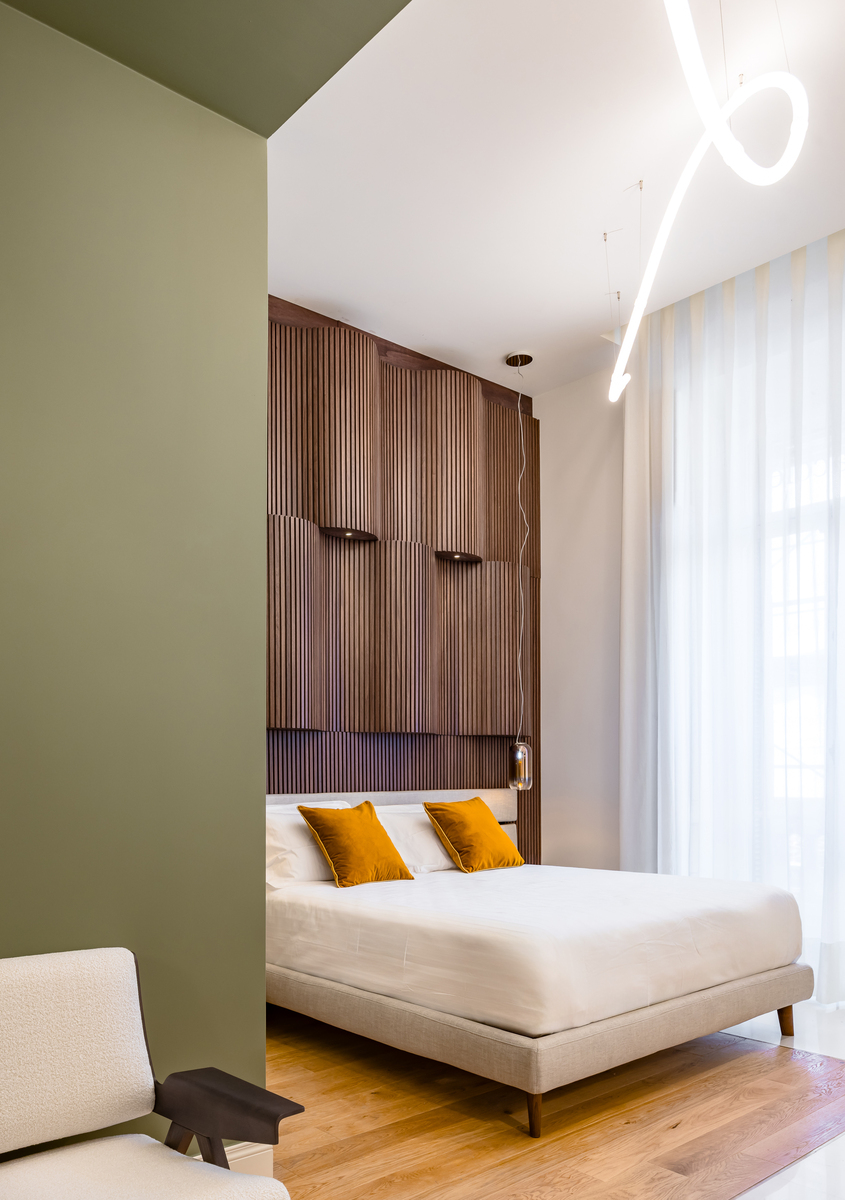 Modern bedroom with olive green wall, wood slat headboard, and Artemide ceiling light in sinuous LED design above a beige bed with mustard cushions.