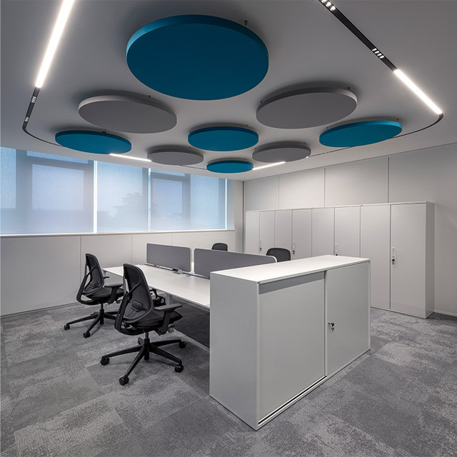 Modern office with circular blue and grey acoustic panels on the ceiling, white modular desks, black ergonomic chairs and diffused and direct-emission lighting.