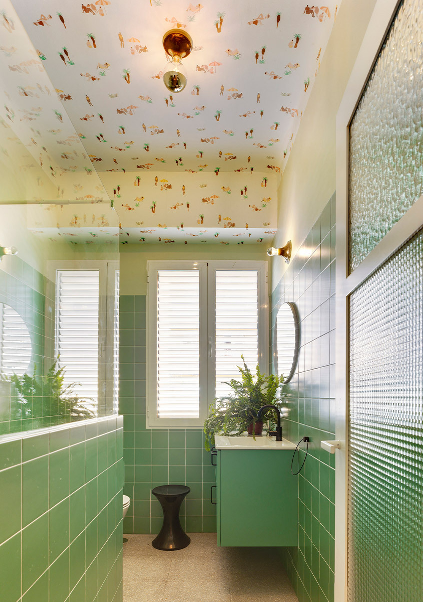 Narrow bathroom with green wall tiles, a wall-mounted sink unit, plants, and a patterned ceiling.
