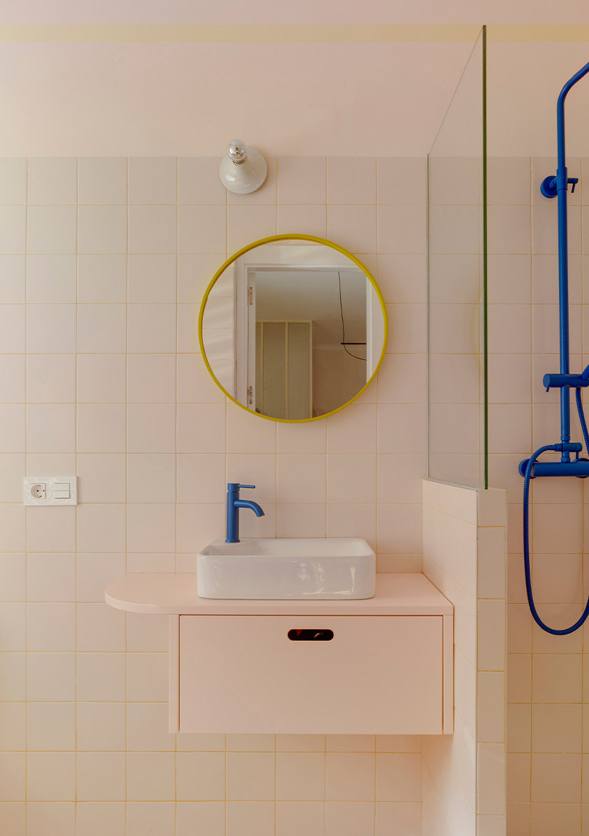 Detail of the bathroom with wall-mounted sink, round mirror with yellow frame, and blue faucet fixtures.