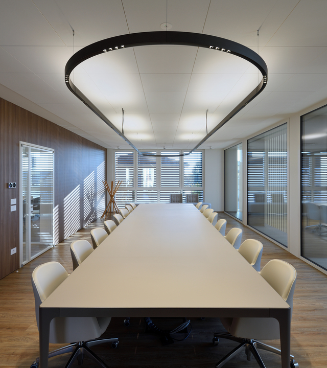 Modern meeting room with glass walls, a large central table and light-colored chairs, illuminated by the A.24 suspended lighting system.