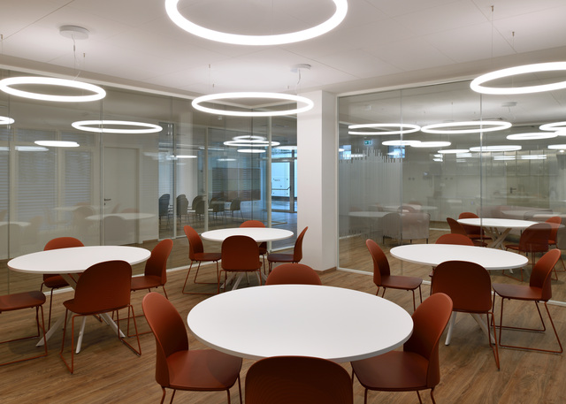 Large room with white round tables and colorful chairs, illuminated by circular Alphabet of Light pendant luminaires.