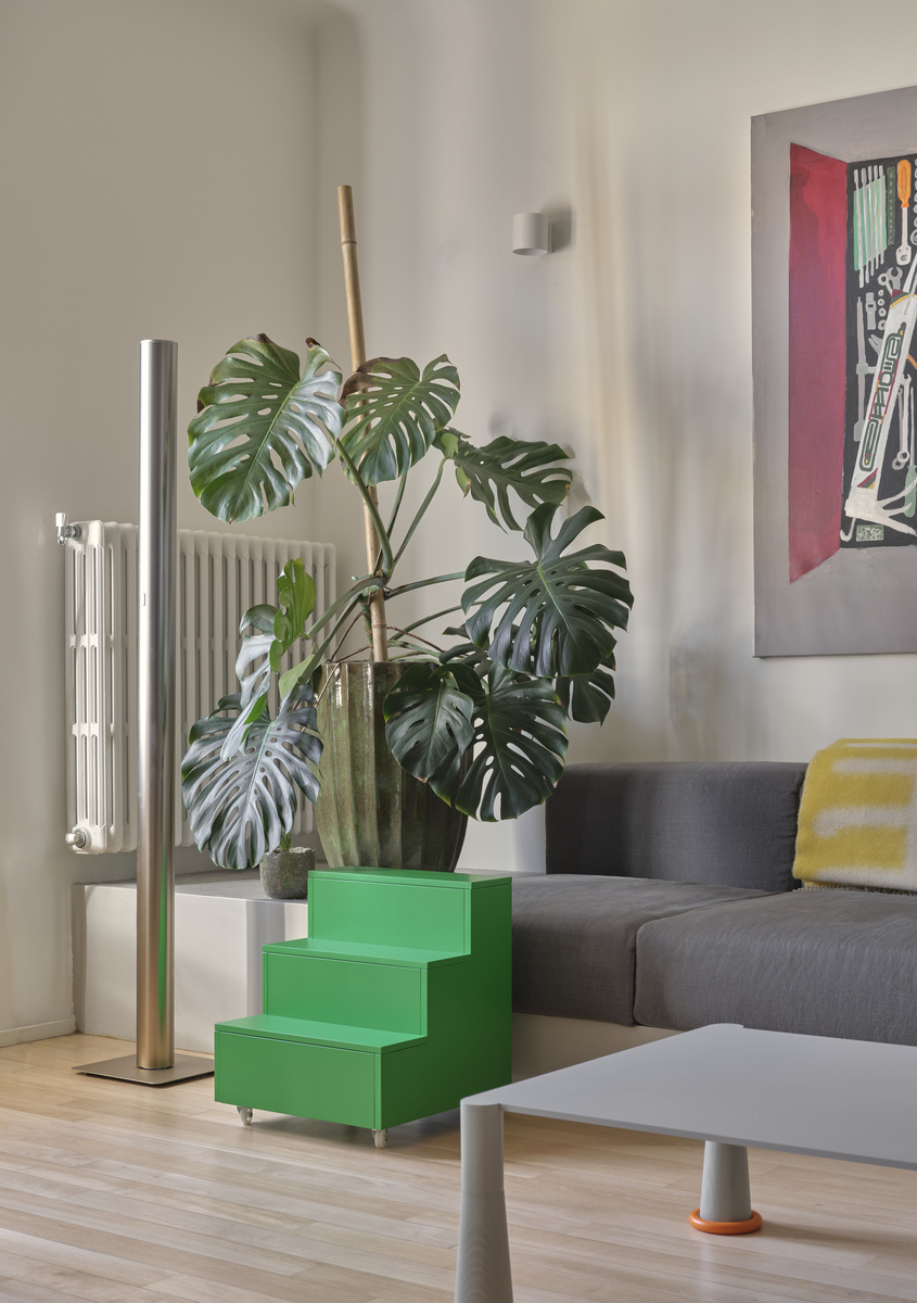 Corner of the living room with Artemide’s Ilio 10 floor lamp, a large potted monstera and a green step-shaped side table beside the sofa.