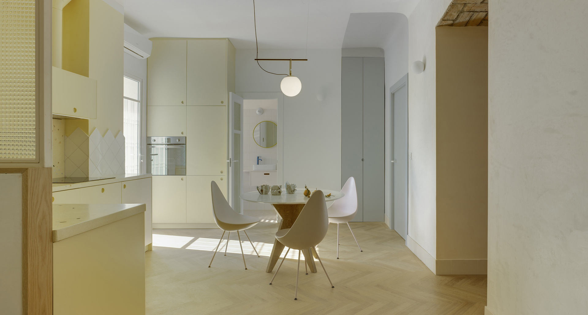 View of the kitchen with dining area, herringbone wood flooring, and cream-colored furnishings, all illuminated by a pendant lamp.