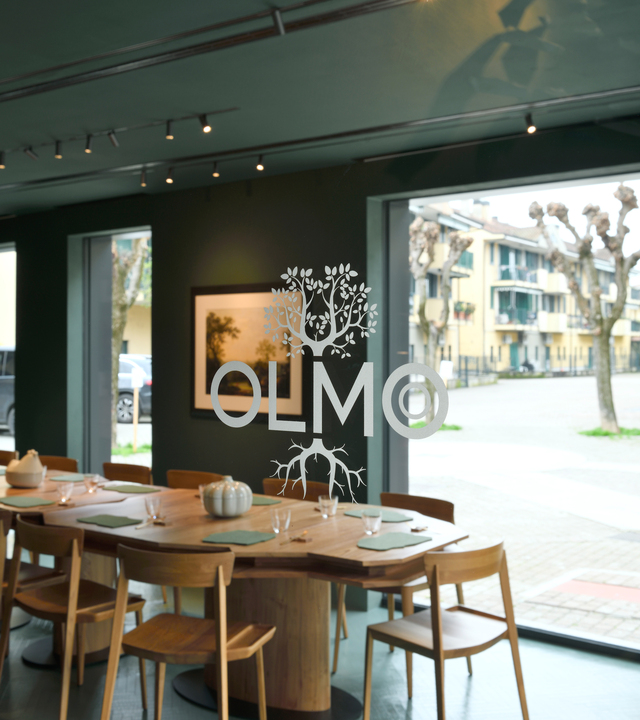 Immagine del logo di Olmo posto su una vetrata del ristorante dove si vede la sala principale.
