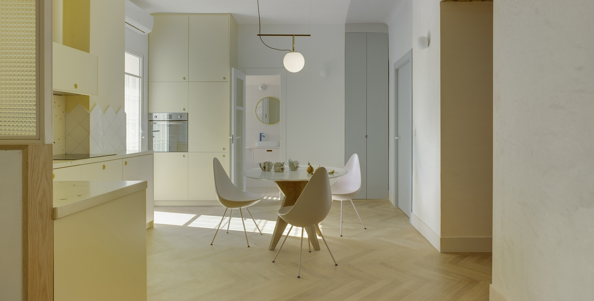 View of the kitchen with dining area, herringbone wood flooring, and cream-colored furnishings, all illuminated by a pendant lamp.