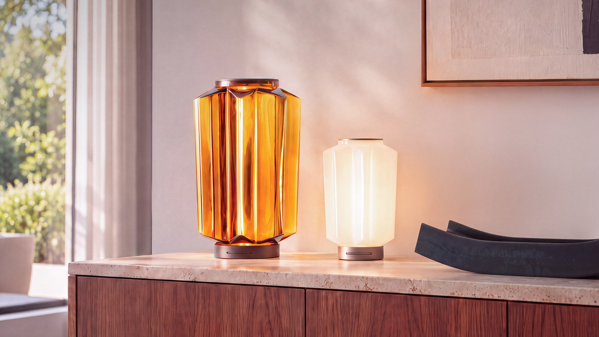 Dos lámparas de mesa Artemide sobre un aparador de madera: izquierda en vidrio ámbar facetado, derecha más pequeña en vidrio opal blanco, ambas con luz cálida.
