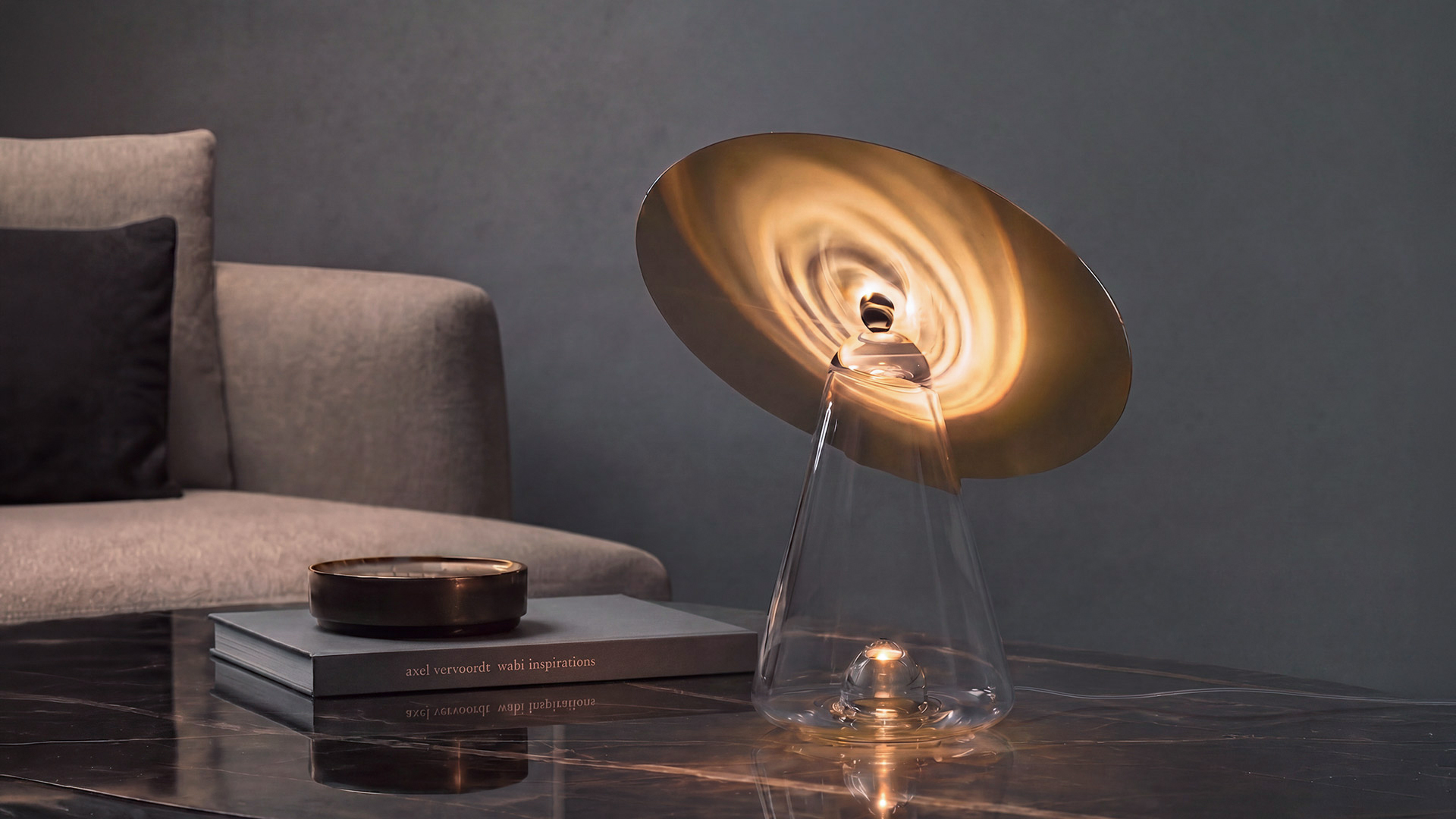Lampe de table Artemide avec base conique en verre transparent et abat-jour disque blanc incliné, allumée sur une table basse en marbre dans un salon moderne.